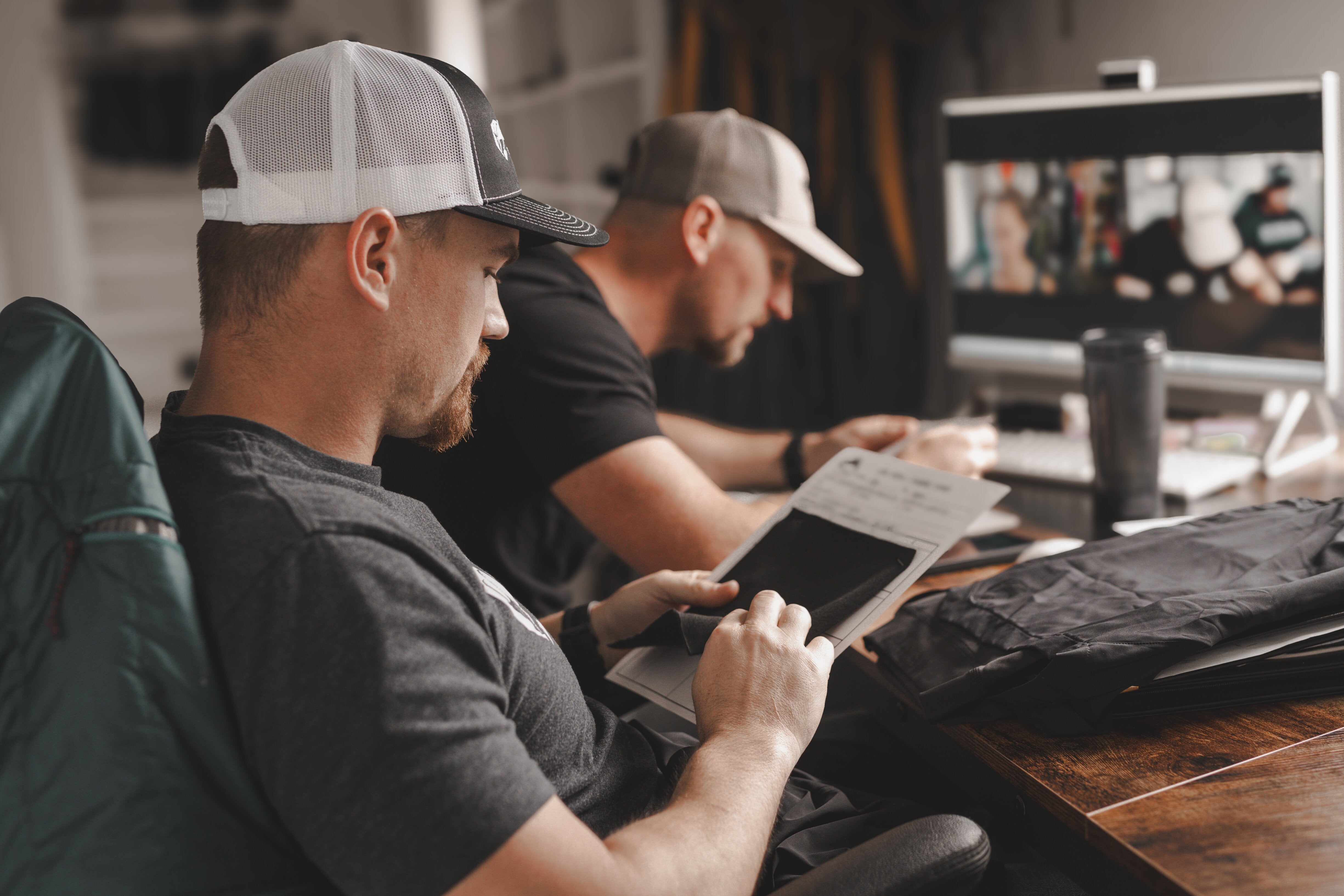 Image of two men wearing ball caps and t-shirts while looking at fabrics and on a computer video call.