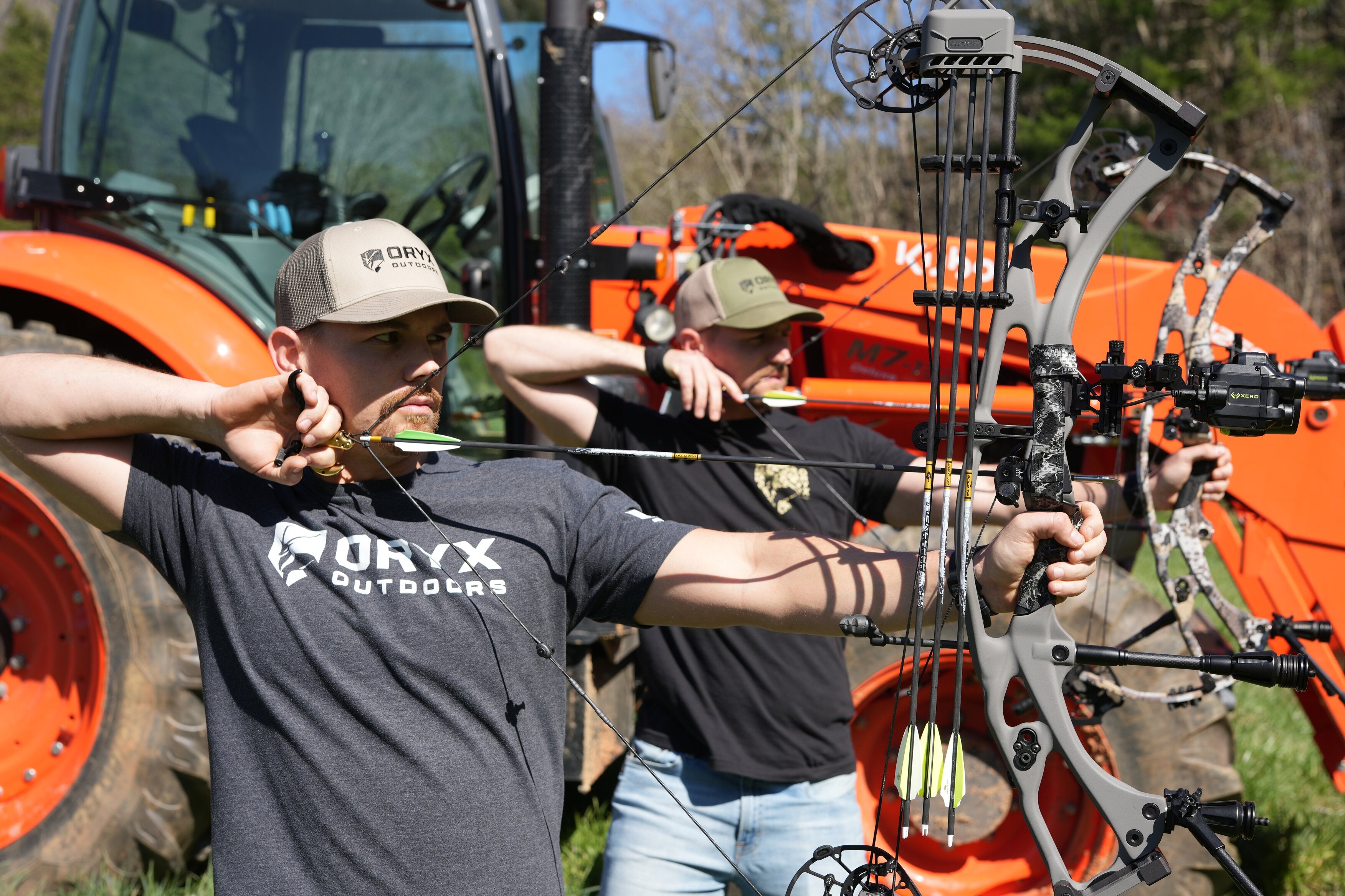 Image of two outdoorsmen with hunting bows aiming at an unseen target while standing beside a large orange tractor.